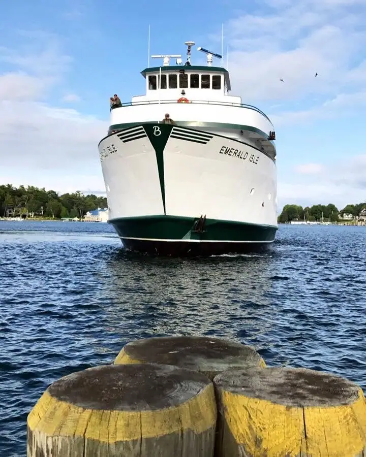 A white-and-green ferry approaches a pier, with three large wooden pilings in the foreground.