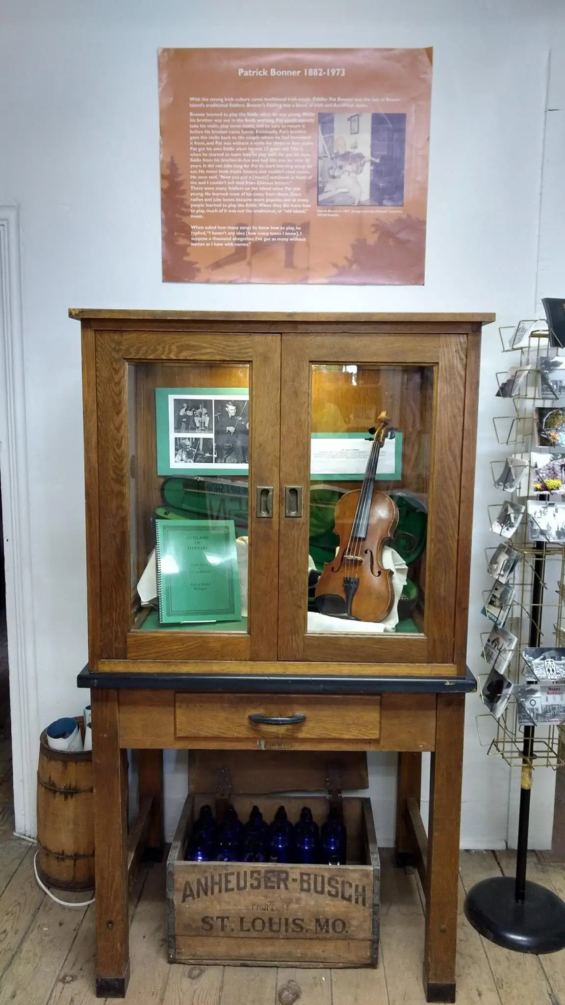 A violin and archival materials are displayed in a wooden glass-front cabinet beneath an informational poster.