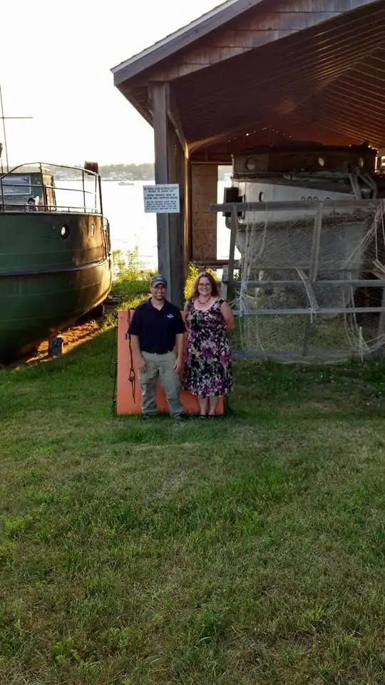 Two adults stand beside boats and a wooden boat shelter near the water.