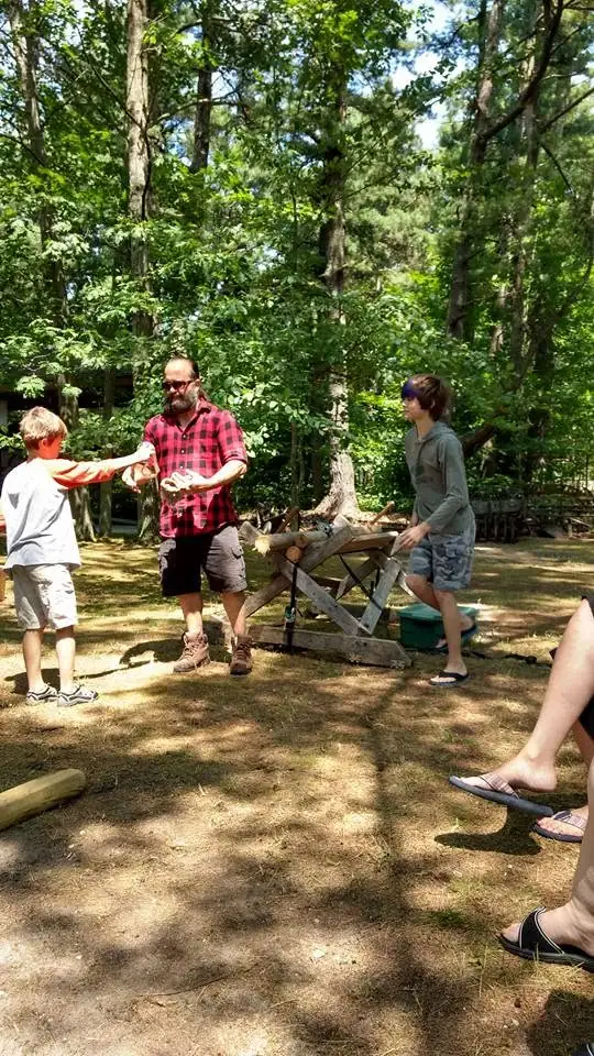 An adult and two children gather around a wooden cart loaded with logs in a wooded clearing.