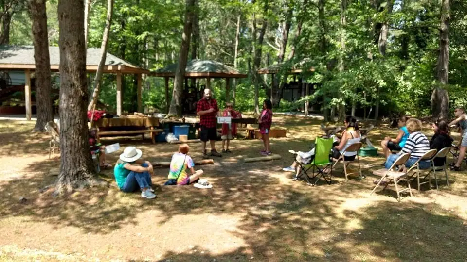 An adult addresses children and seated adults in a wooded clearing.