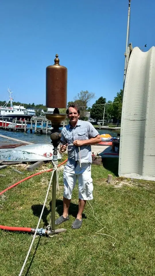 A man stands beside a tall bronze whistle and rope near boats and a dock.