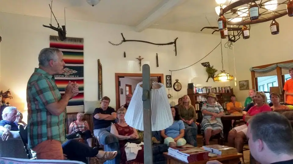 A man speaks to a seated audience in a room decorated with books, bottles, textiles, and mounted objects.