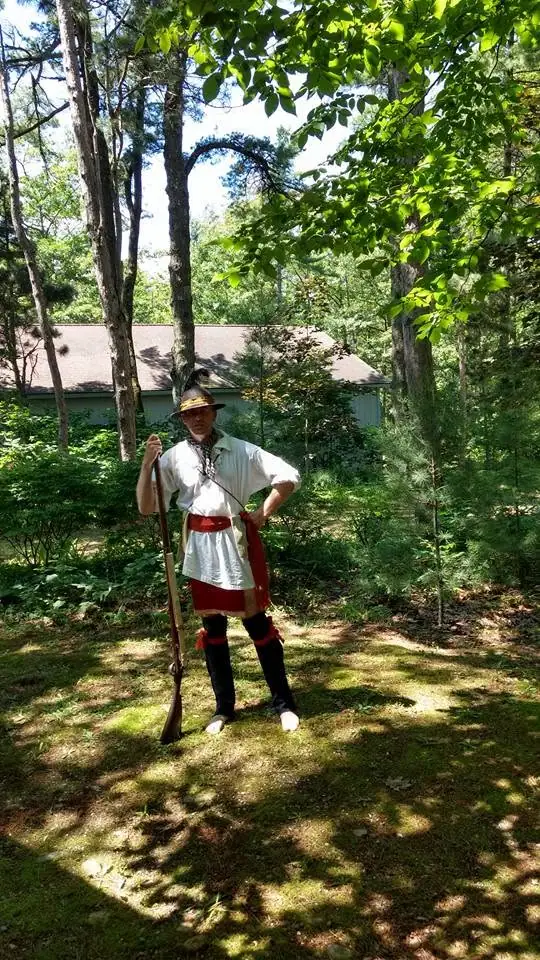 A person in a white and red costume stands with a long firearm among trees beside a low green building.