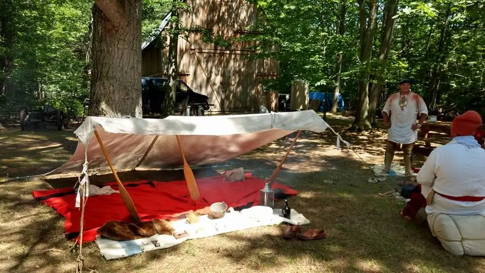 People in costume gather around a canvas shelter and displayed utensils in a wooded clearing.