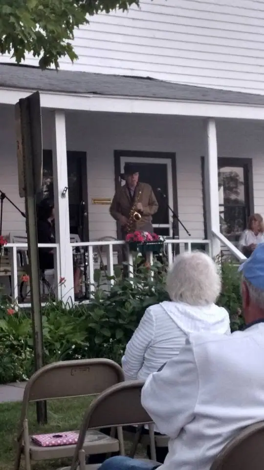 A saxophonist performs on a porch as several people watch from folding chairs.