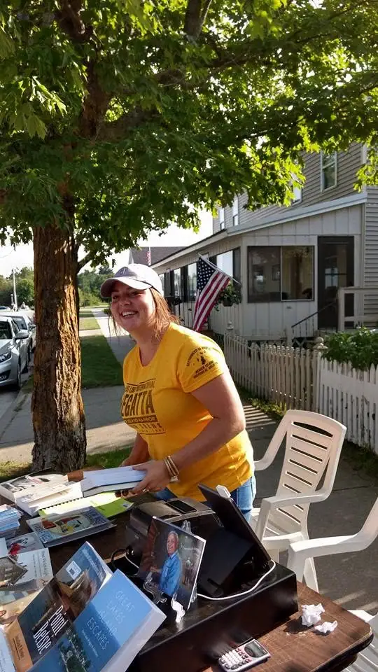 A woman in a yellow shirt stands beside a table of brochures outside a house.