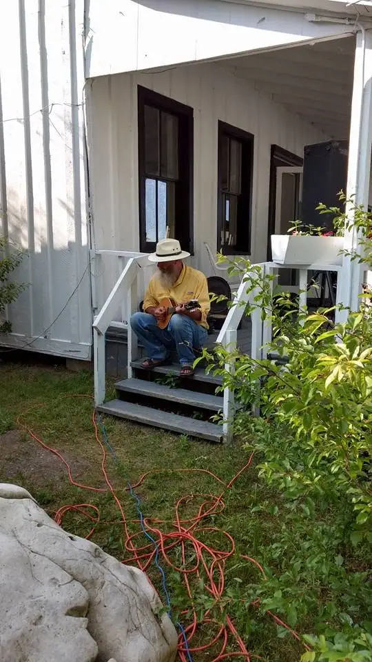 A bearded man in a hat plays a small stringed instrument on the steps of a white porch.