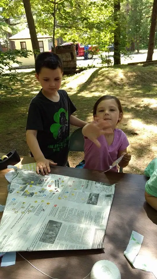 Two children gather around an open newspaper at an outdoor table beneath trees.