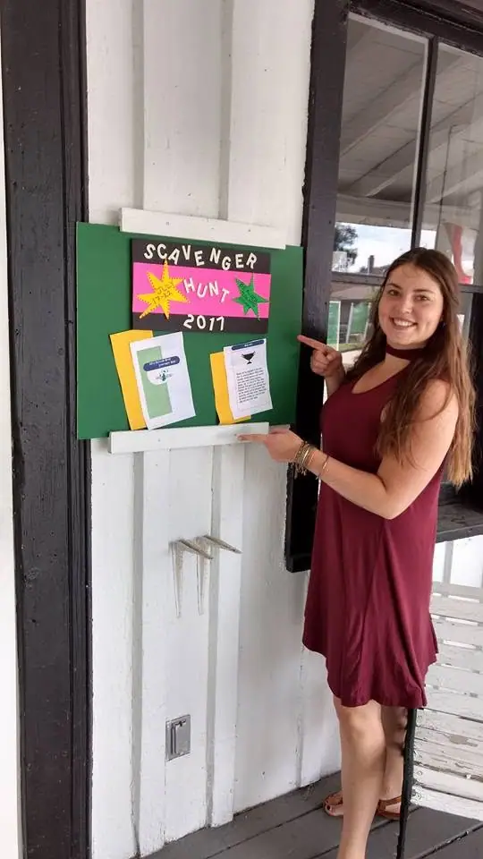 A woman points to a green bulletin board advertising a 2017 scavenger hunt.