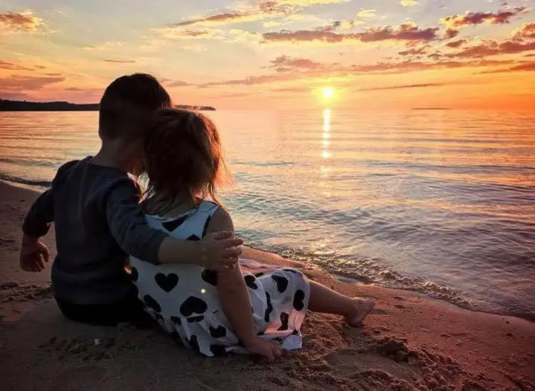 Two children sit together on a sandy shoreline, watching the sun set over calm water.