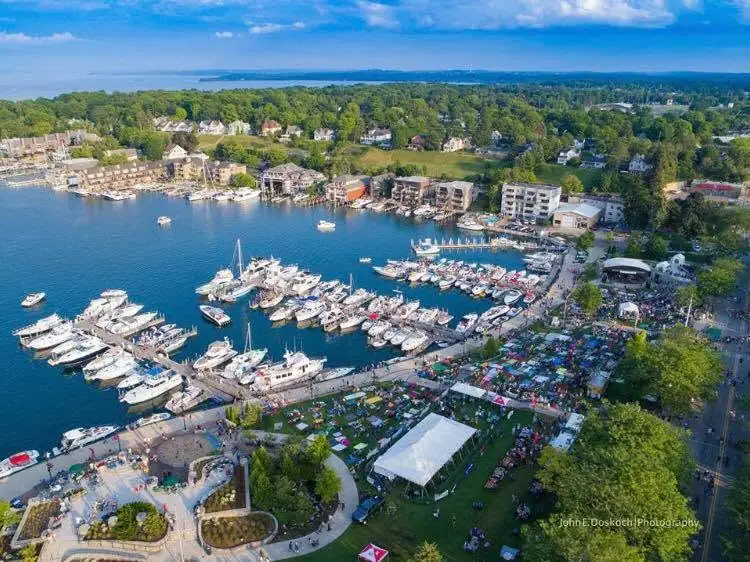Aerial view of a marina crowded with boats, waterfront buildings, docks, and an outdoor gathering beneath a large white tent.