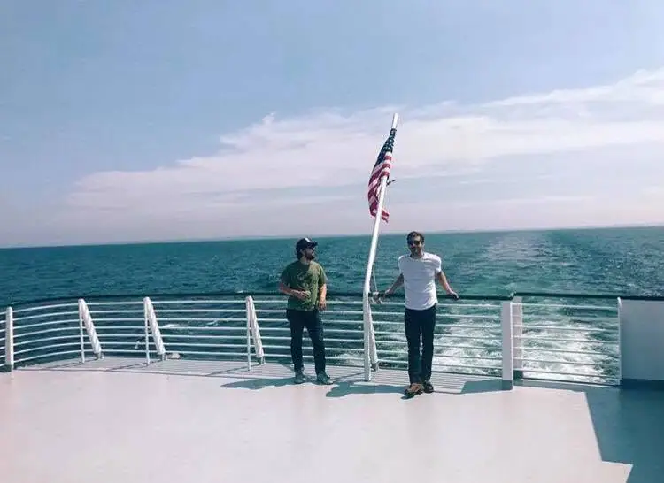 Two men stand on the open deck of a ferry beside an American flag, with a wake trailing across the water.