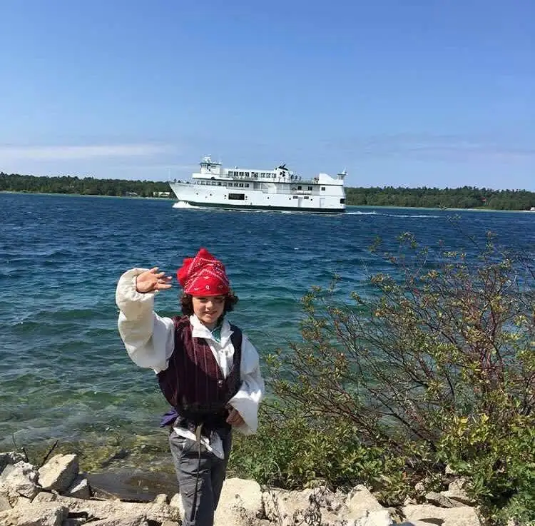 A costumed person waves beside a rocky shoreline as a white ferry passes across the water.