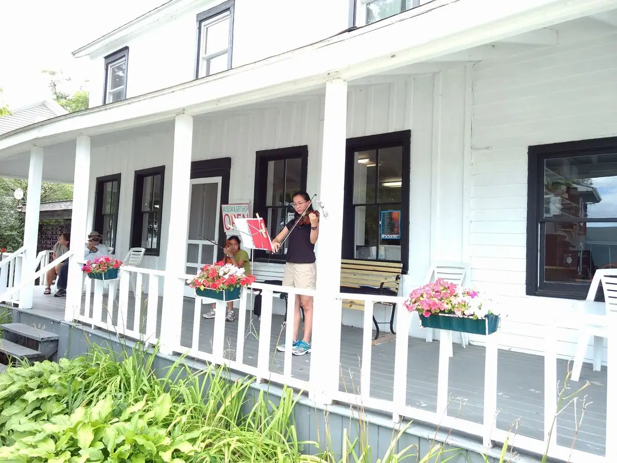 Two musicians perform on the porch of a white wooden building beside flower boxes.