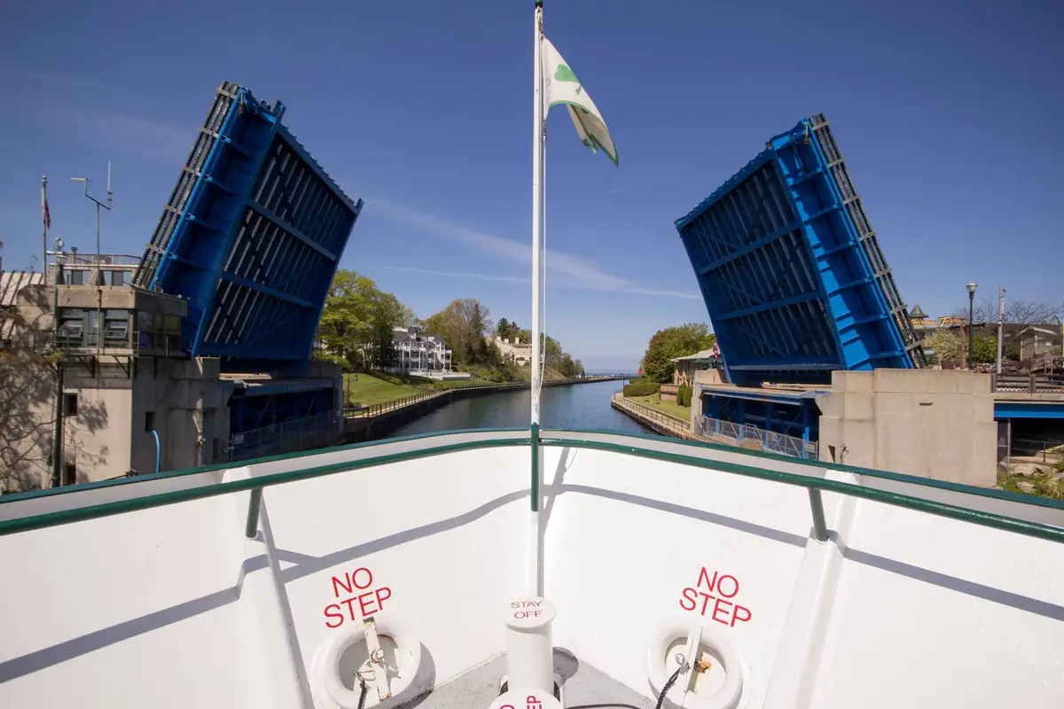 A boat approaches a raised blue drawbridge, with a green-and-white flag and safety lettering on the bow.