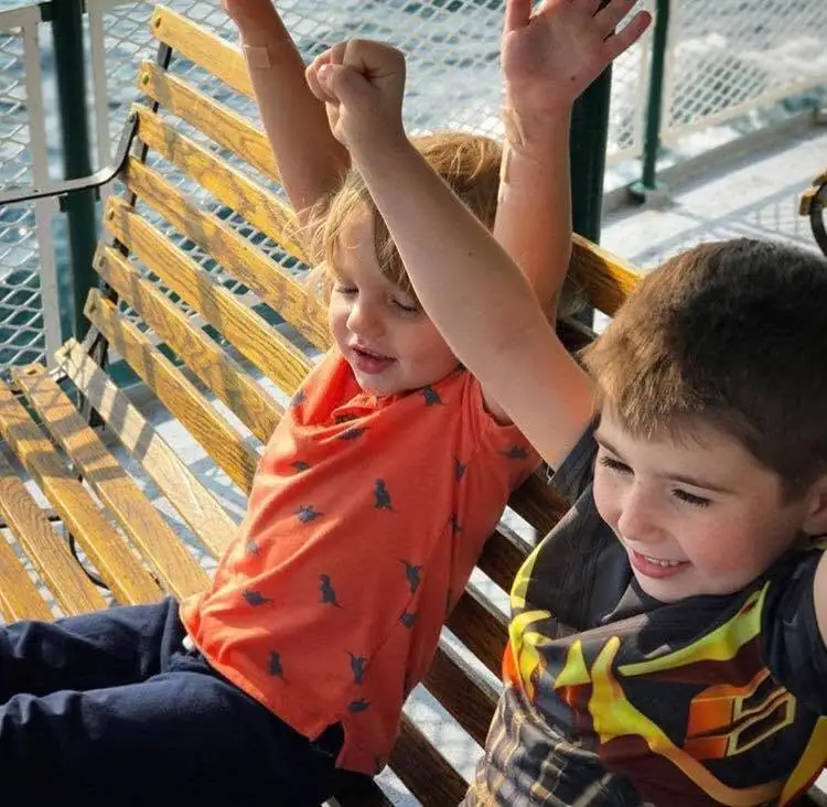 Two young children sit on a wooden slatted bench with their arms raised beside a metal safety railing.