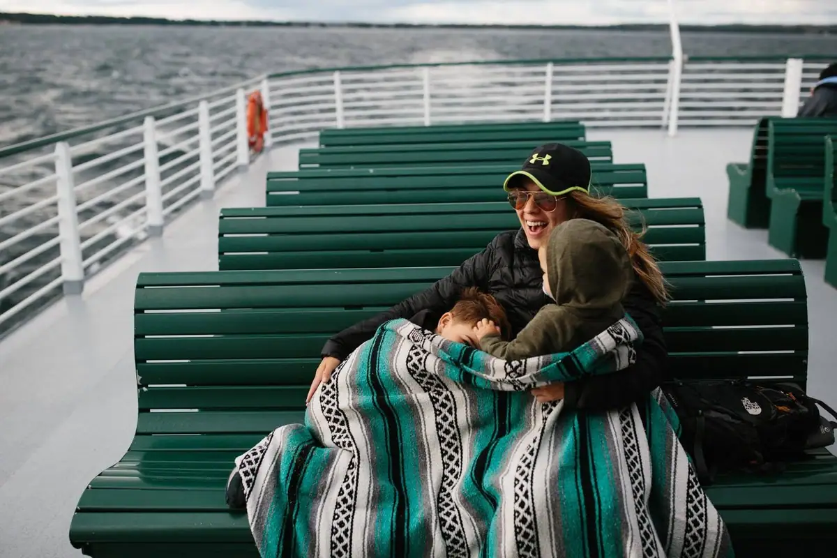 A woman sits with two children under a patterned blanket on a ferry’s open deck.