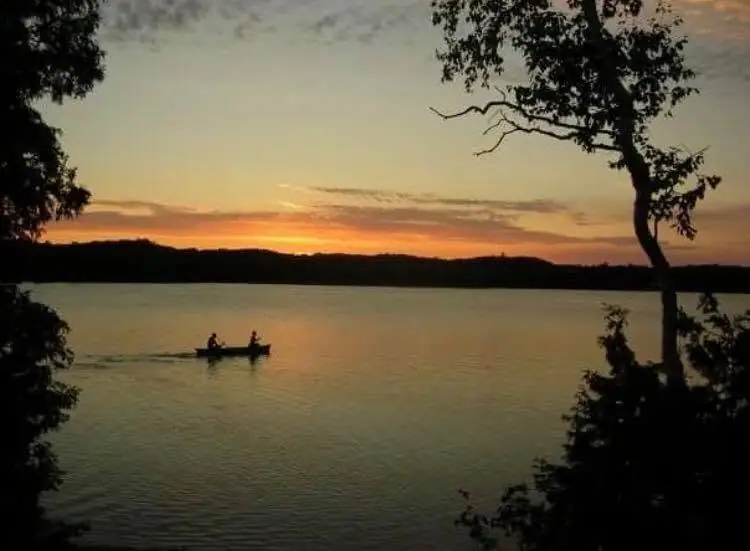 Two people in a canoe cross calm water at sunset, framed by silhouetted trees.