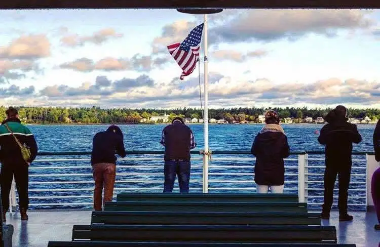 People stand on a passenger boat deck beside benches, looking toward a wooded shoreline beneath an American flag.