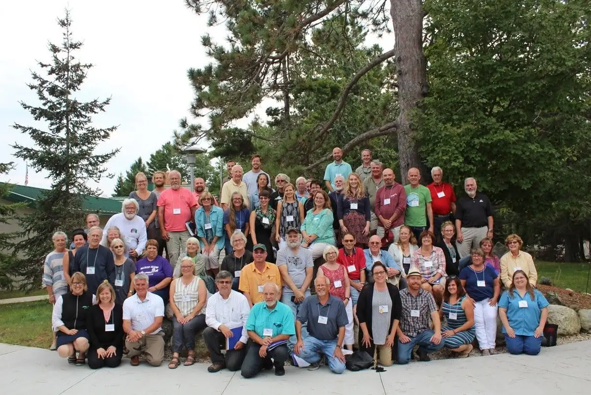 A large group of adults poses outdoors among trees, many wearing name badges.