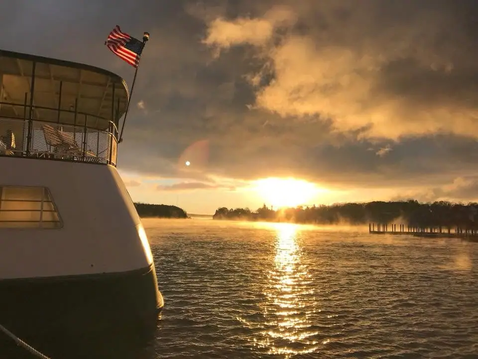 A passenger vessel with an American flag sits on rippling water at sunset beside a pier.