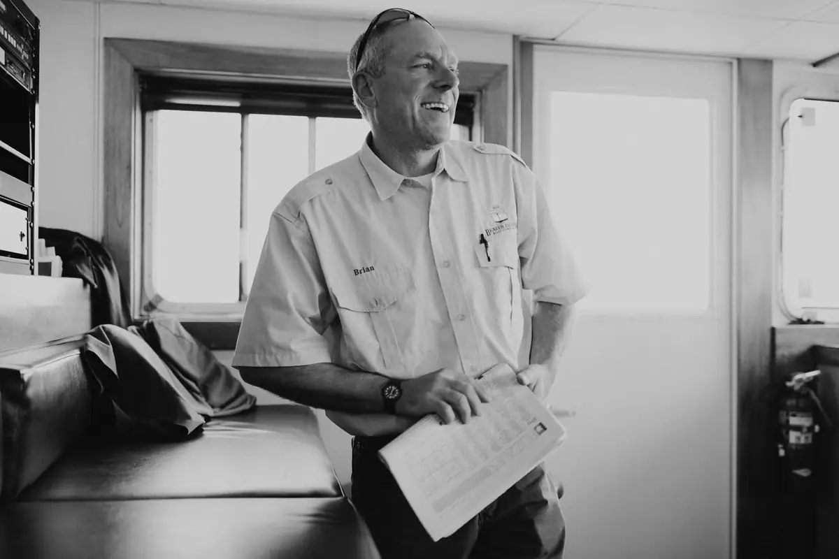 A smiling uniformed man holds a clipboard inside a boat cabin.
