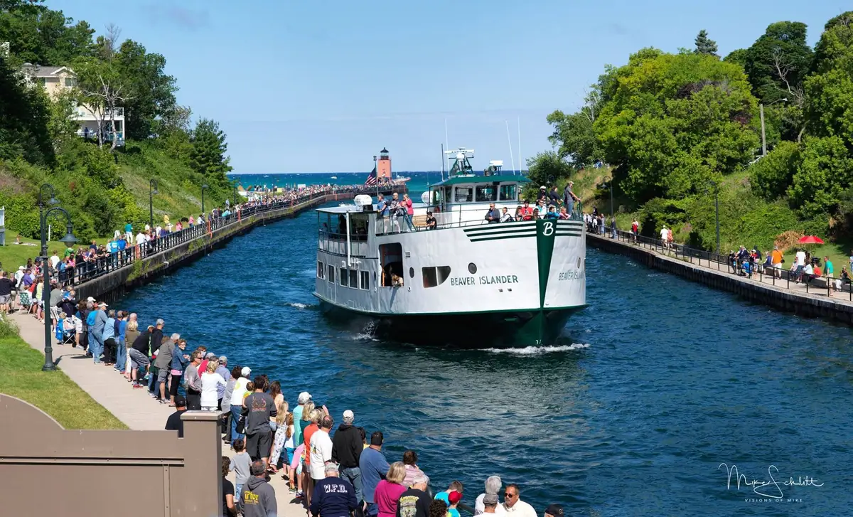 A white-and-green ferry passes between crowded walkways toward a red lighthouse.