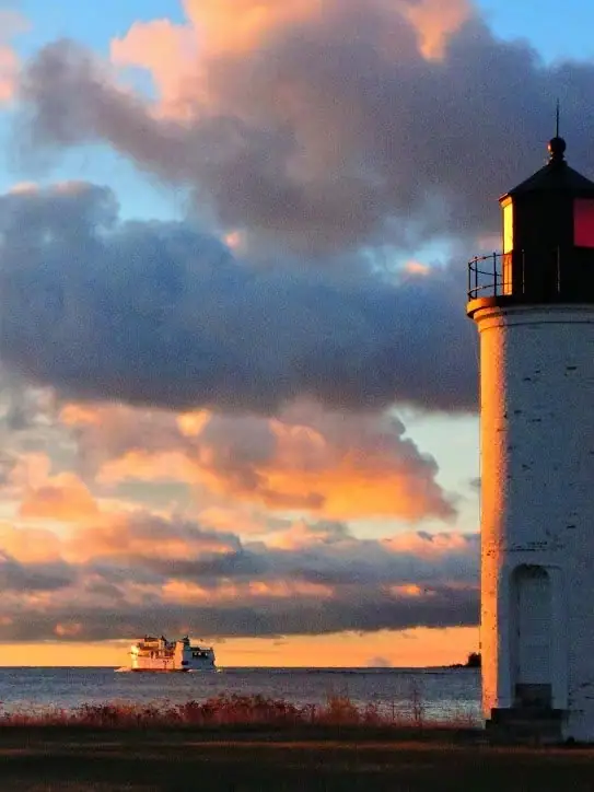 A white lighthouse and distant ferry stand against a dramatic sunset sky over calm water.
