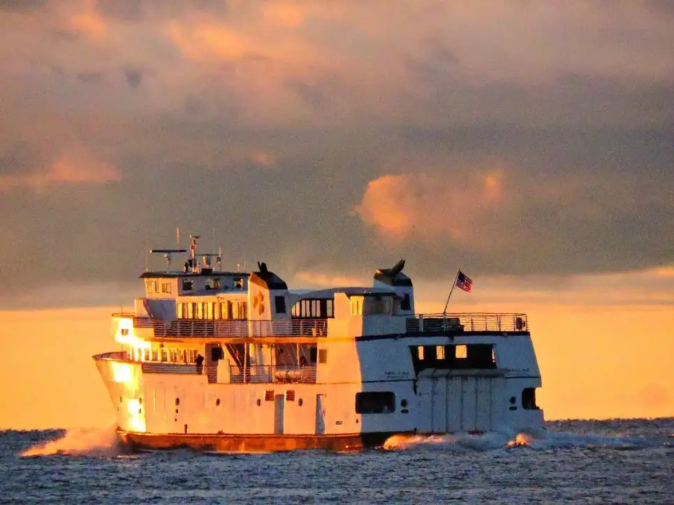 A large white ferry travels across dark water beneath orange-lit clouds at sunset, flying an American flag.