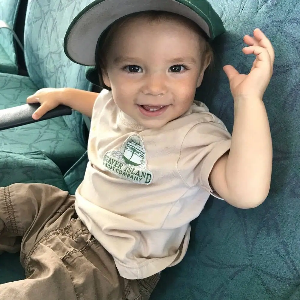 Smiling young child in a green cap and boat-company shirt seated on a patterned teal couch.