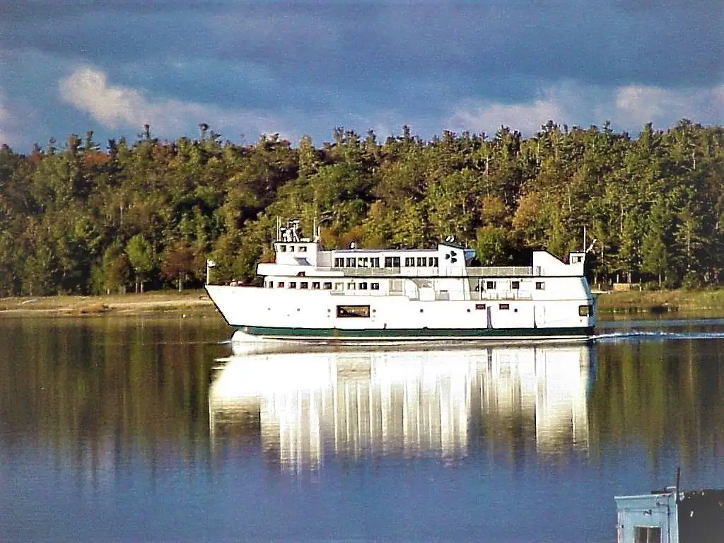 A white multi-deck ferry crosses calm water beside a forested shoreline, with its reflection below.