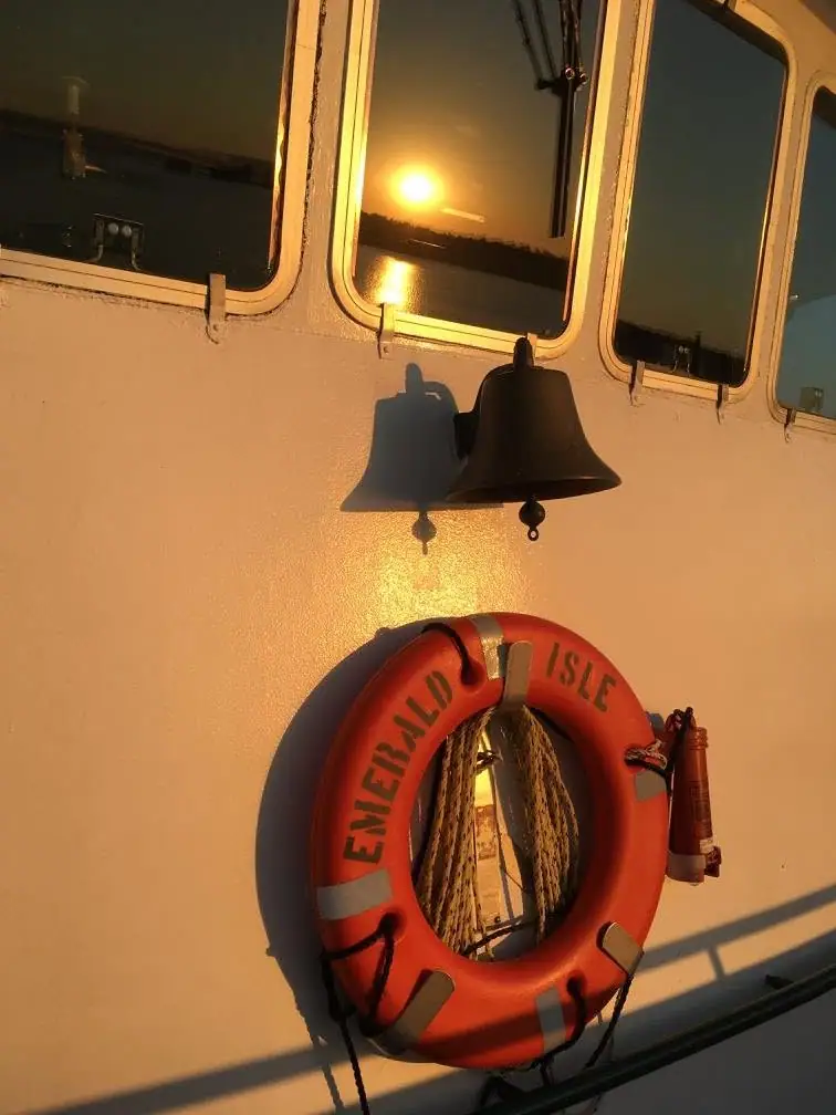 A ship's exterior with a bell, an orange lifebuoy marked “EMERALD ISLE,” and a low sun reflected in the windows.