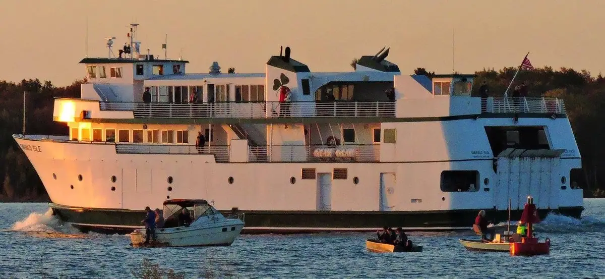 A large white ferry passes several small boats on rippling water in warm evening light.