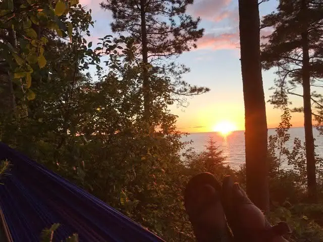 Sunset over calm water viewed through trees from a hammock, with a person's feet in the foreground.