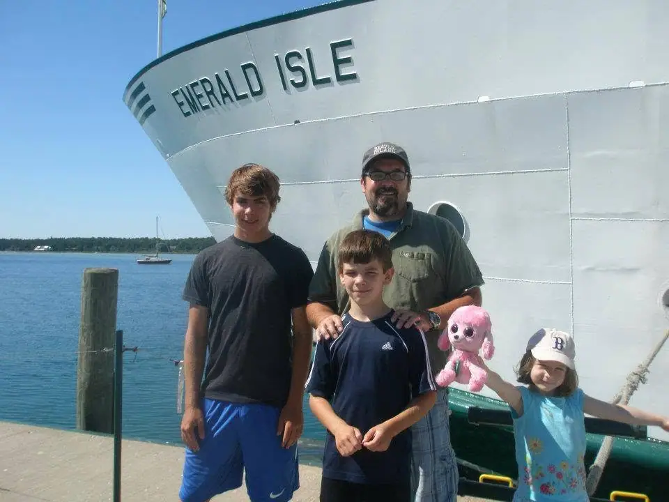 Four people pose beside the bow of a large ferry at a dock, with a small sailboat in the background.