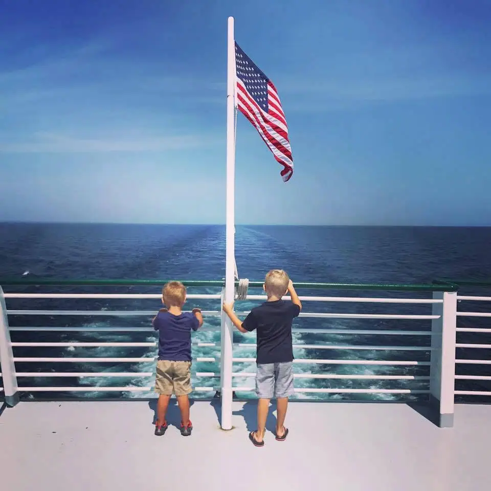 Two boys stand at a stern railing beneath an American flag as a vessel leaves a foamy wake.