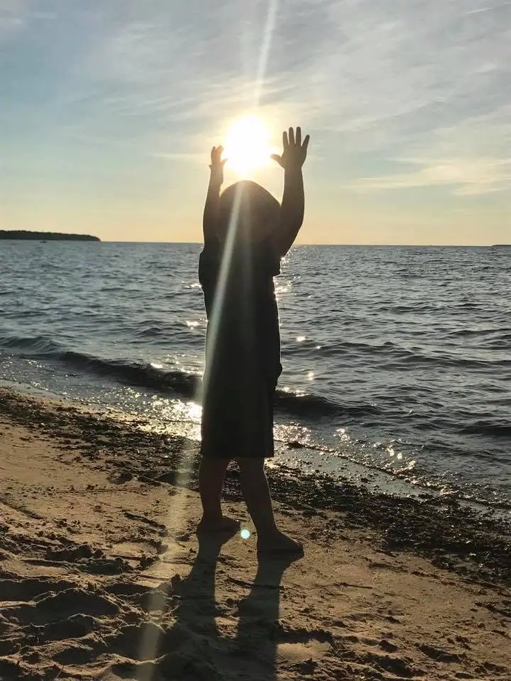 Backlit person standing on a sandy shoreline with both arms raised toward the low sun.