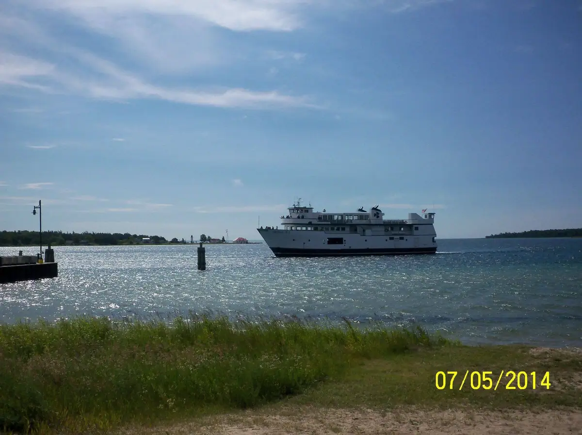 A white passenger ferry crosses bright water beside a grassy shoreline, with a pier and channel marker in view.