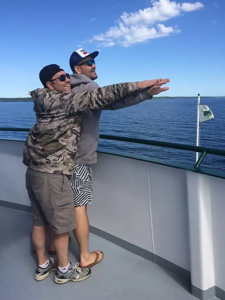 Two men stand together on a vessel deck beside open water under a bright, partly cloudy sky.