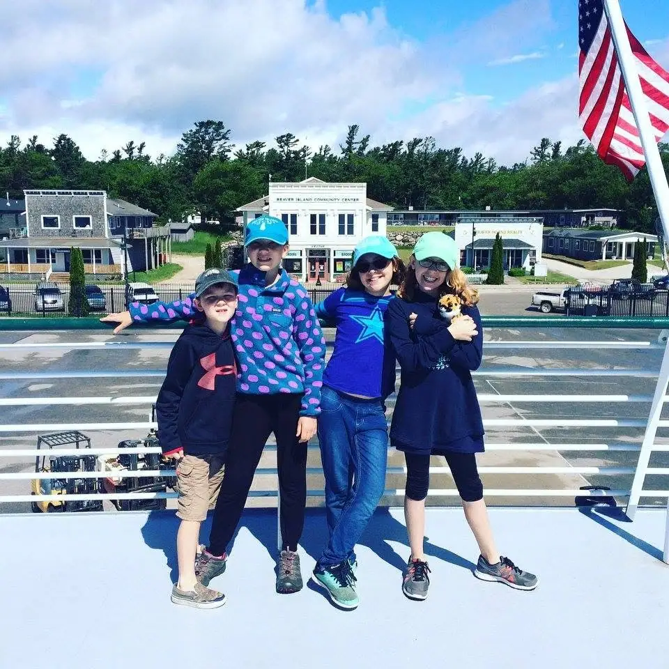Four children pose on a ferry deck with a small dog, an American flag, and buildings across the street.