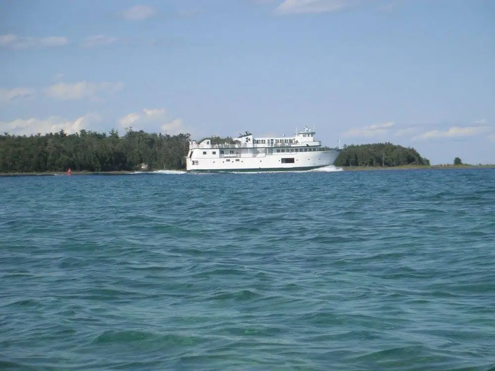 A white passenger ferry travels across rippling water beside a forested shoreline under a partly cloudy sky.