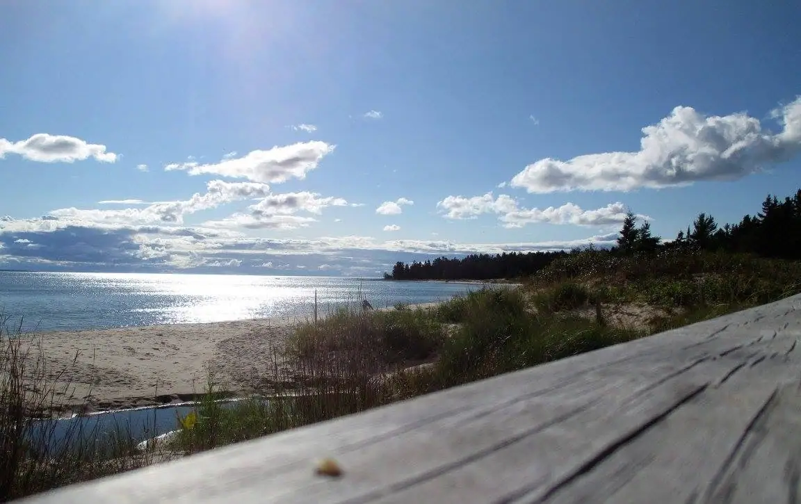 A sandy shoreline and calm water viewed past grasses and a weathered wooden deck beneath a cloud-filled sky.