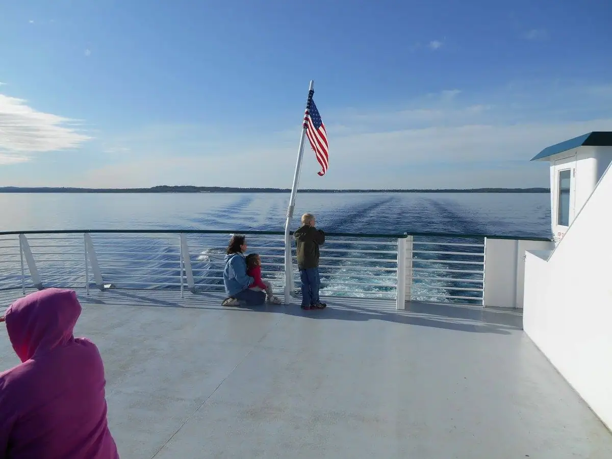 Passengers on a ferry deck watch the wake beneath an American flag.