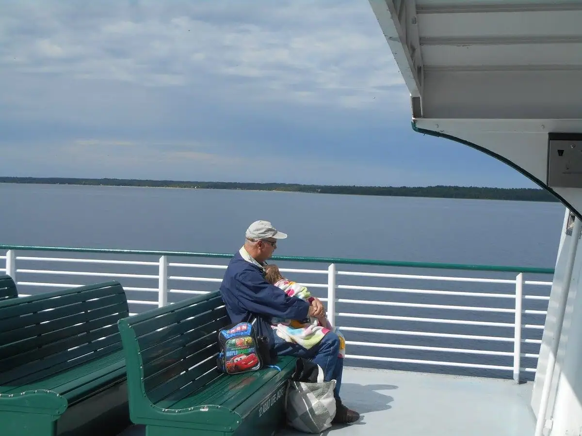 A man holds a blanket-wrapped child on a ferry deck beside green benches and a calm expanse of water.