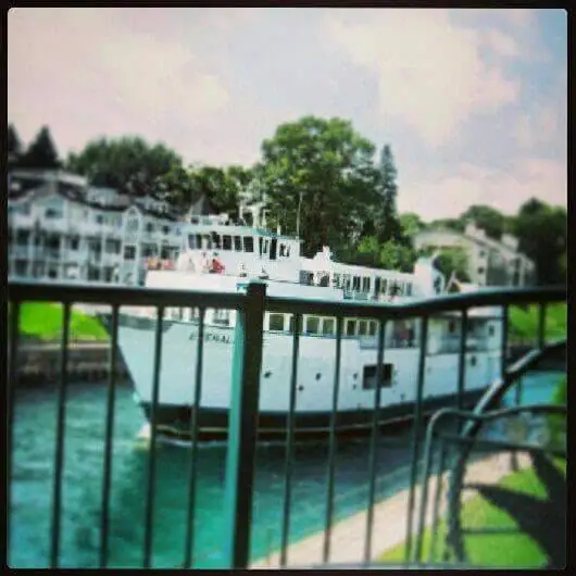 A white ferry passes a waterfront railing, with buildings and trees along the shore behind it.