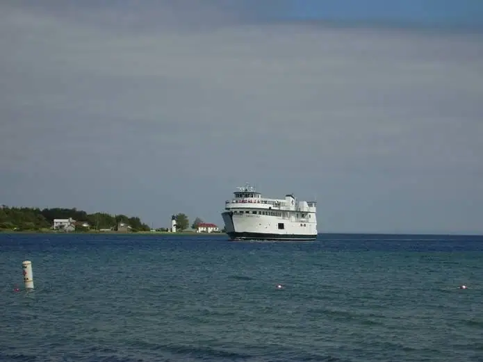 A white passenger ferry travels across blue water with a wooded shoreline and buildings behind it.