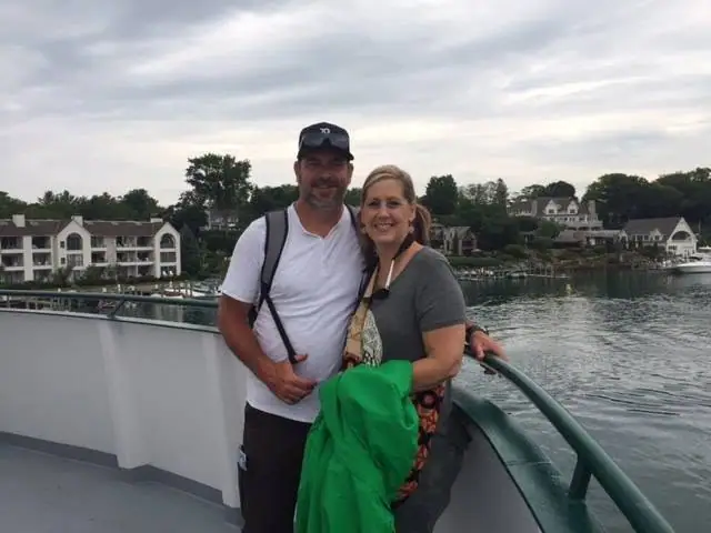 Two smiling adults stand beside a green railing on a boat deck with waterfront homes behind them.