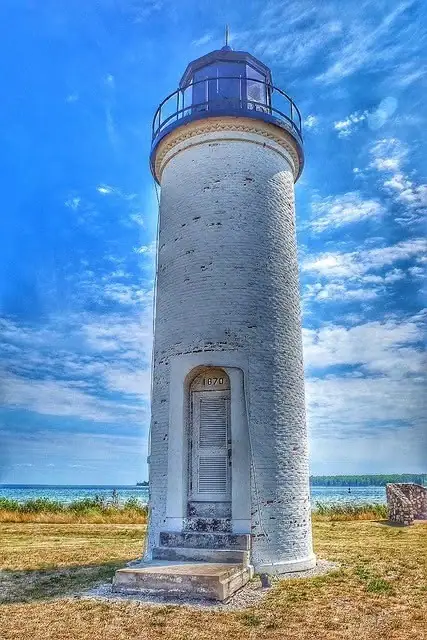 A white cylindrical lighthouse with a black lantern room stands beside a grassy shoreline beneath a partly cloudy sky.