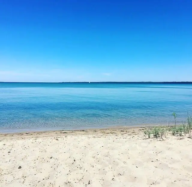 A sandy beach borders calm blue water beneath a clear sky, with a distant shoreline on the horizon.